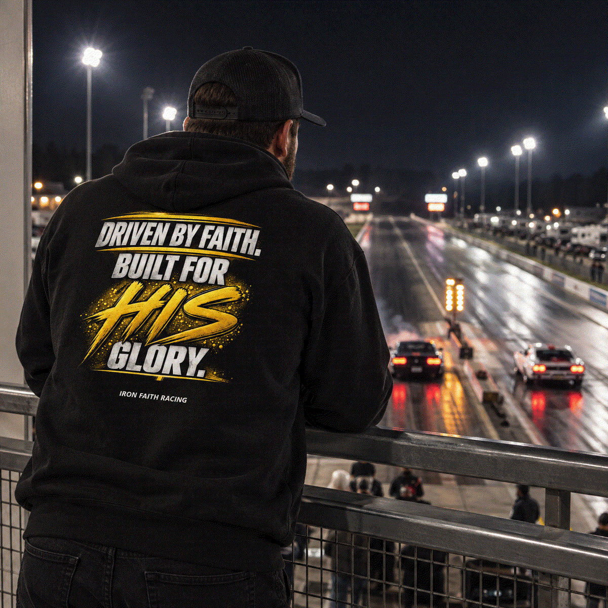 Man in Faith Hoodie at the track rail at night watching cars under the lights