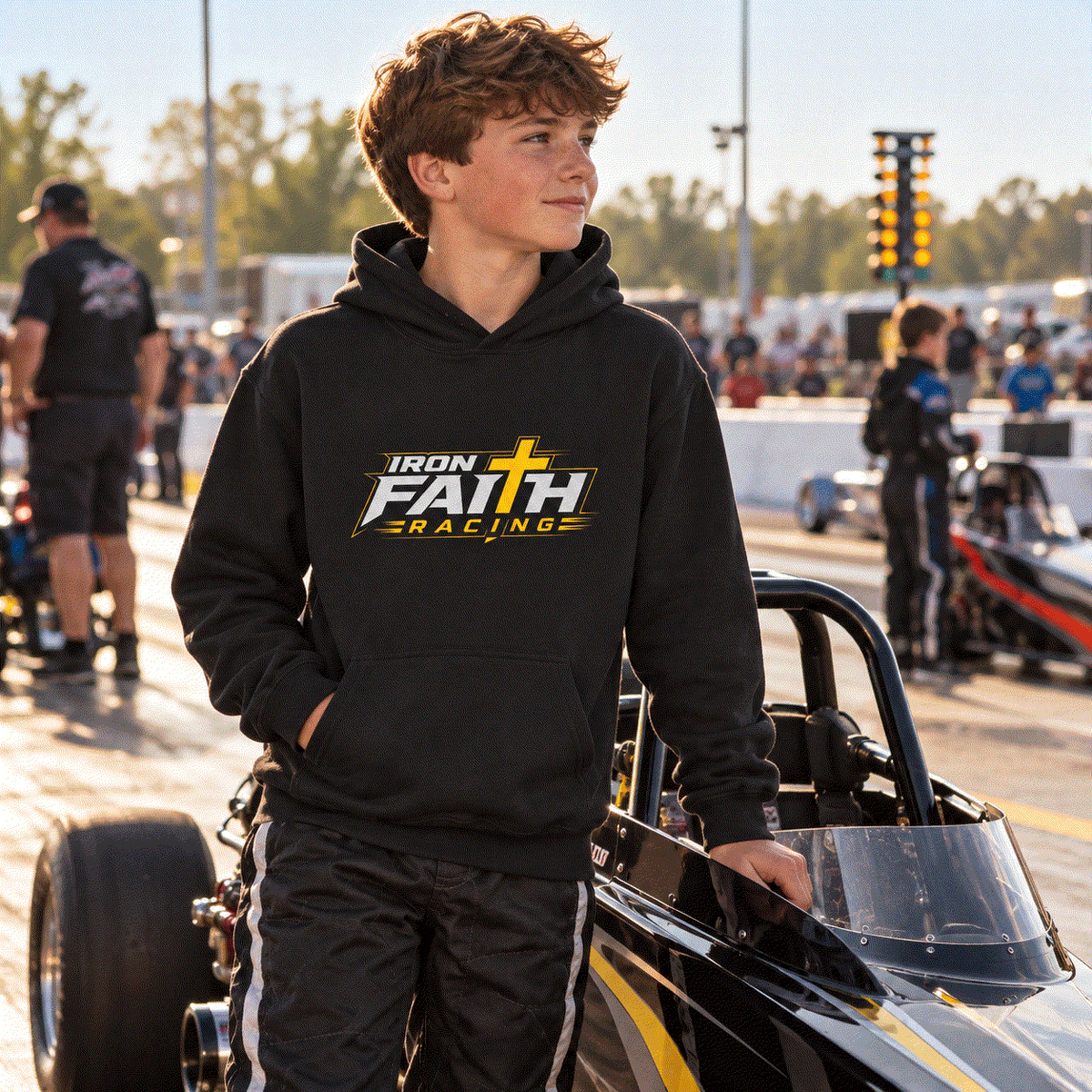 Boy in Iron Faith Racing youth hoodie standing by a junior dragster in staging